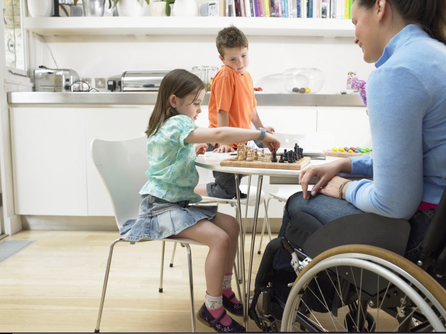 A woman in a wheelchair and two children play chess at a table in a bright, modern kitchen. The girl is making a move while the boy stands and watches, smiling.