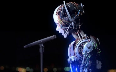 A humanoid robot with exposed wires and circuitry is singing or speaking into a microphone against a dark, blurred background.