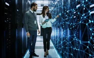 Two people stand in a server room with rows of computer racks. One person holds a tablet while the other looks on. A digital network visualization overlays the scene, suggesting data or network connections.