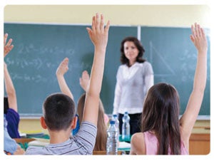 Kids in the classroom with one hand up in front of a female teacher