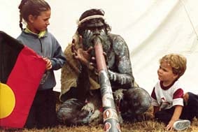 Aboriginal man playing didgeridoo with young girl holding the Aboriginal flag and little boy looking on