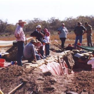 Doctor Judith Field stands in a trench addressing 11 individuals kneeling and standing around the outside.