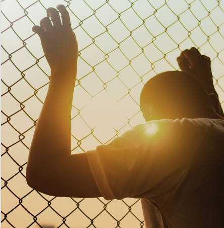 a young person is standing behind a detention fence. They are looking at the setting sun.