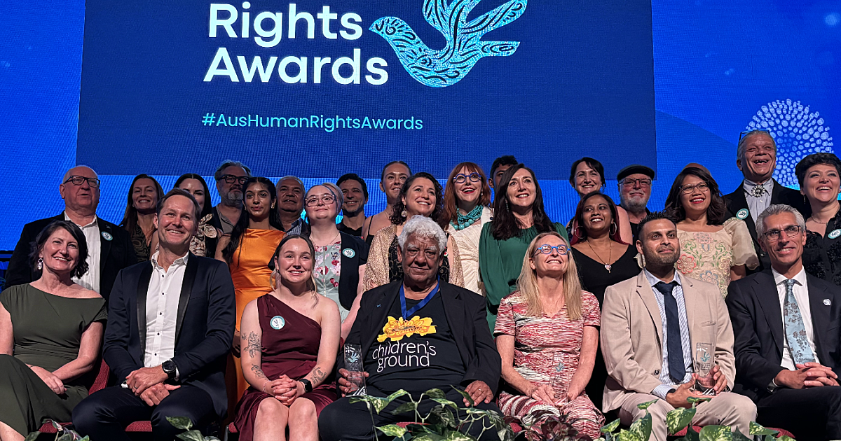 people sitting in a row of seats with many people standing behind them. in the background is a sign which says Human Rights Awards 2025