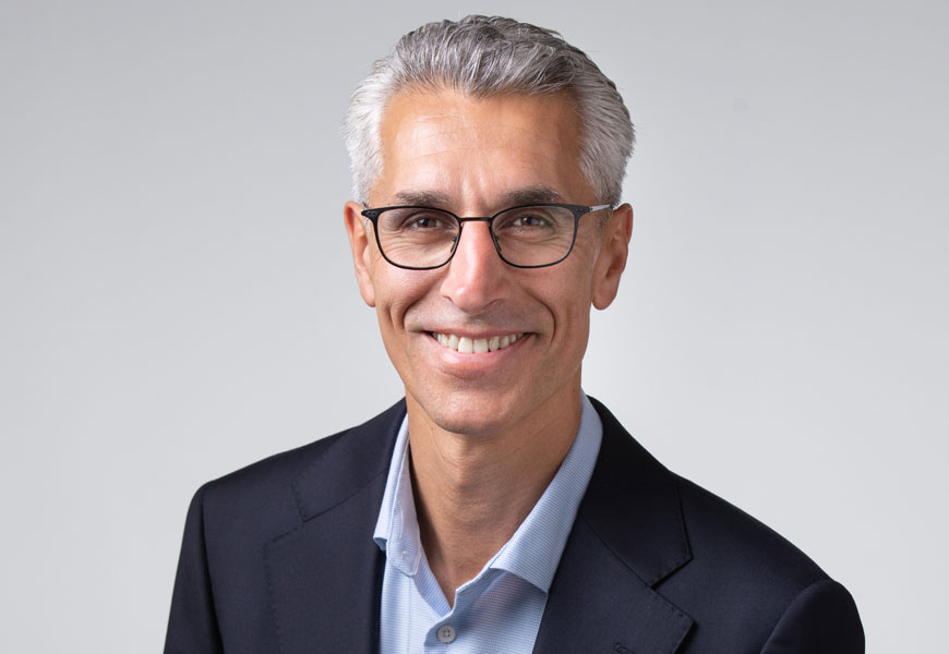 A middle-aged man with short grey hair and glasses, wearing a dark blazer over a light blue shirt, smiles at the camera against a plain light grey background.