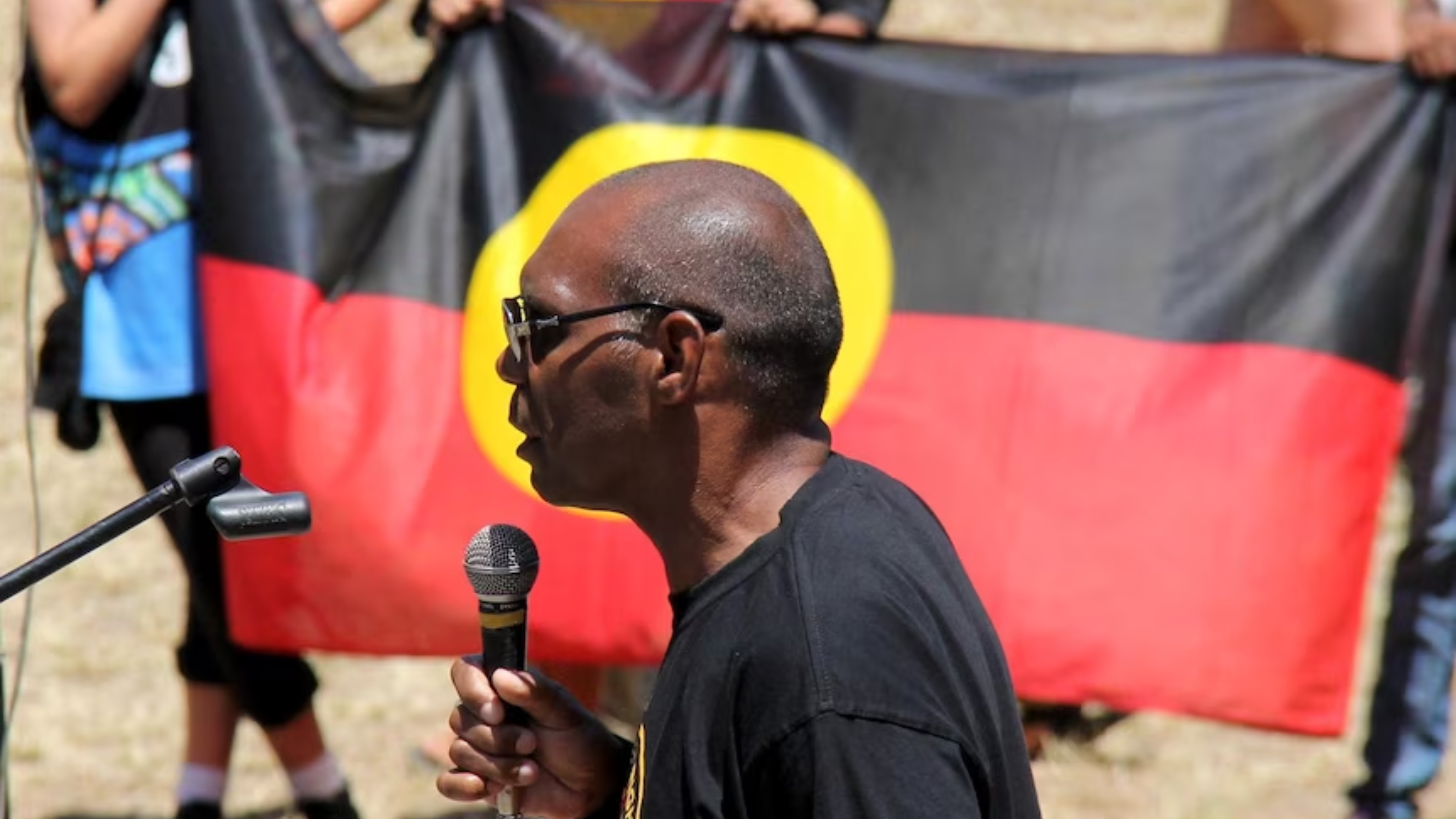 A First Peoples man is standing in front of an Aboriginal flag and is holding a microphone and speaking to a corwd ofmpeople.