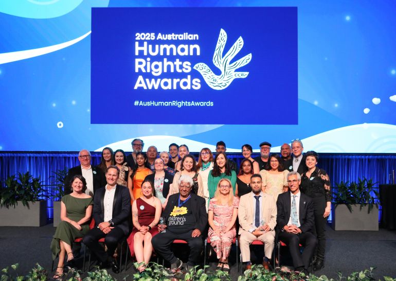 A diverse group of people pose together onstage in front of a large 2025 Australian Human Rights Awards sign, featuring a stylised dove. Some are seated whilst others stand behind, all smiling. The stage is decorated with plants.