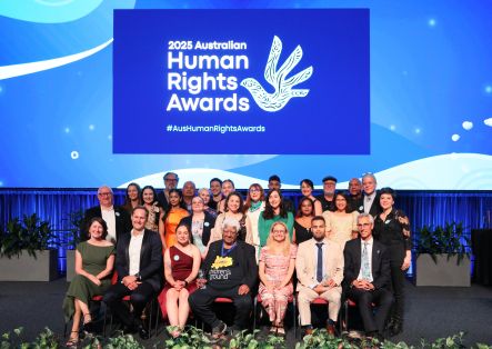 A diverse group of people pose together onstage in front of a large 2025 Australian Human Rights Awards sign, featuring a stylised dove. Some are seated whilst others stand behind, all smiling. The stage is decorated with plants.