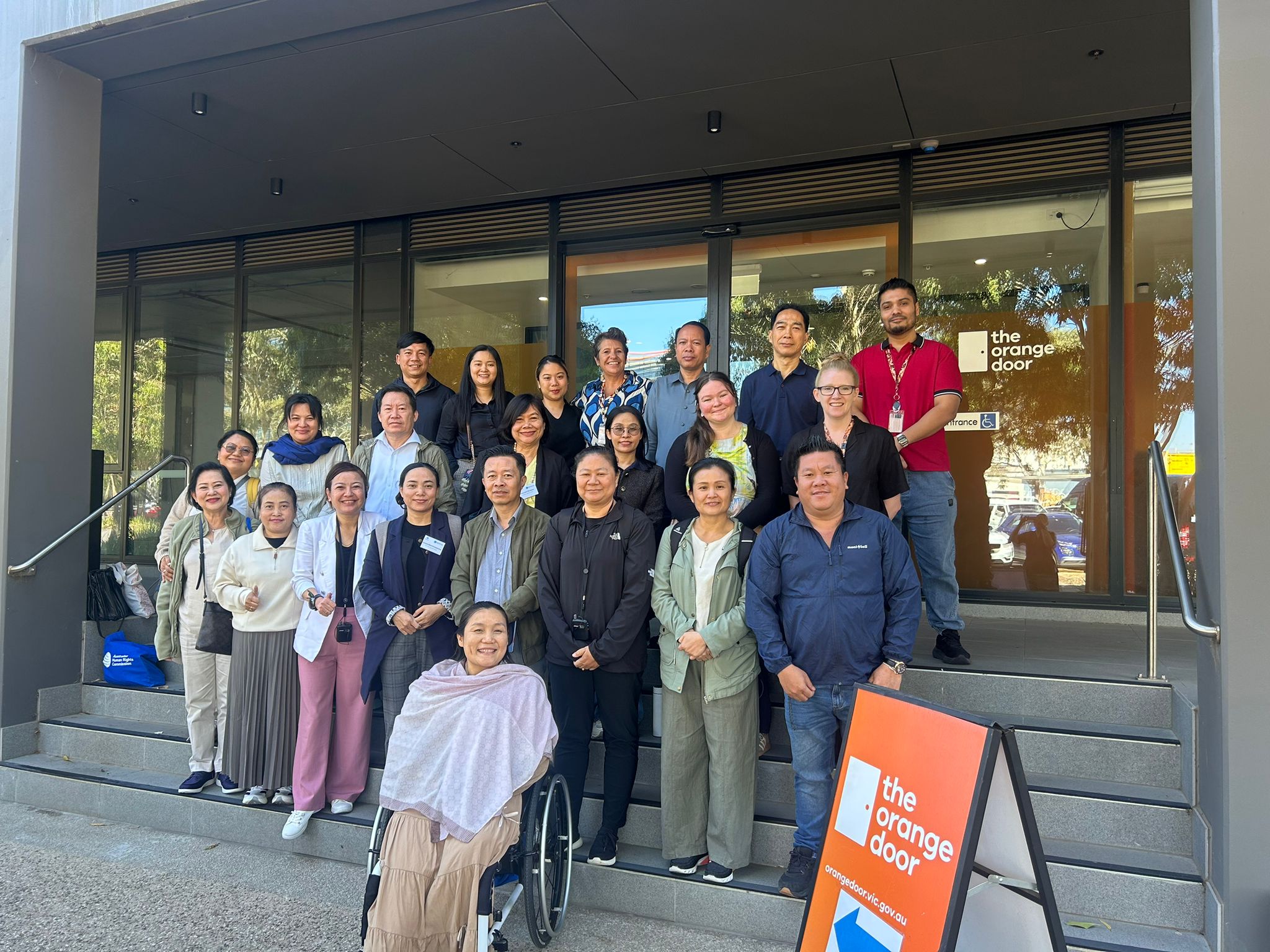 Group of people standing on steps smiling in camera. The sun is bright and the people are standing in front of The Orange Door, a domestic violence shelter in Melbourne, Victoria.