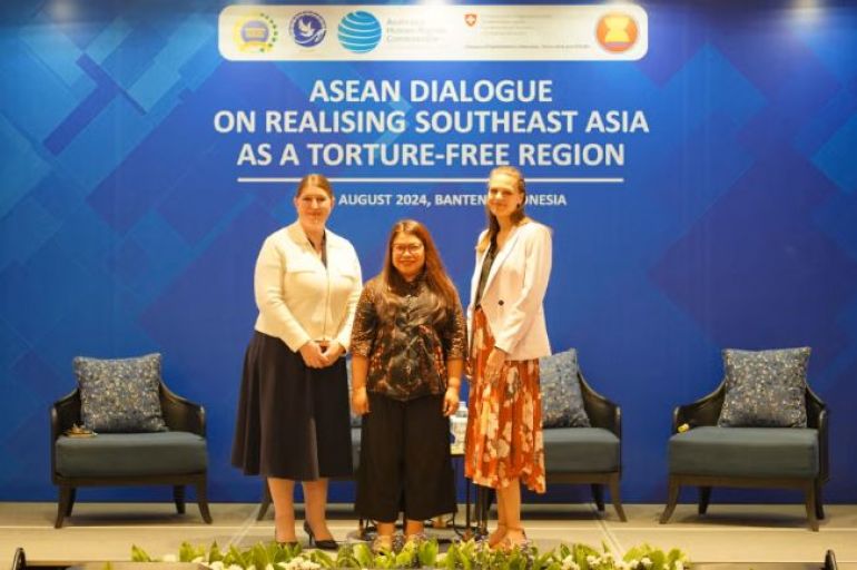 Three women stand on a stage in front of a blue backdrop that reads ASEAN Dialogue on Realising Southeast Asia as a Torture-Free Region, with two empty chairs and a small table beside them.