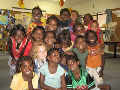 A group of smiling children, mostly with dark hair and skin, pose together indoors in a classroom, some hugging each other and looking at the camera. Art, chairs, and classroom decorations are visible in the background.