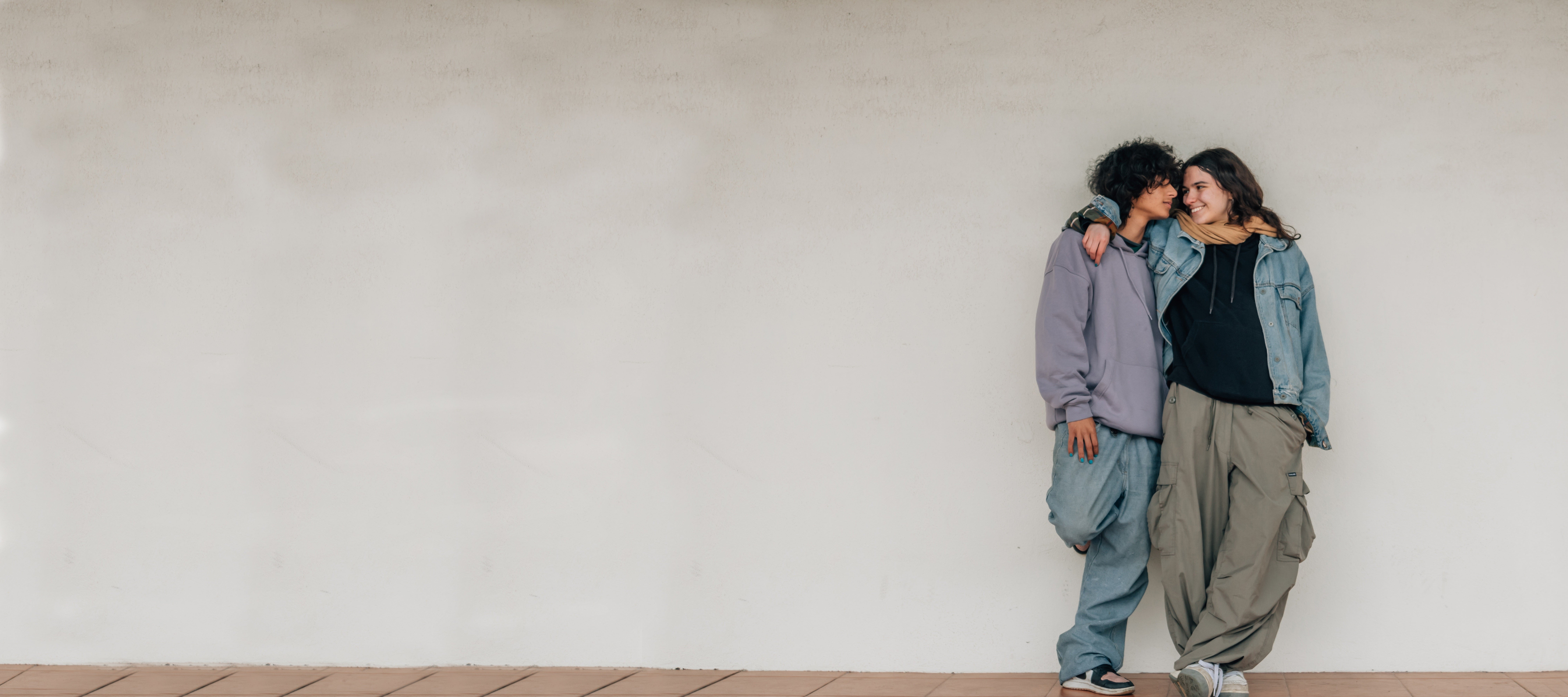 Two young people stand close together against a plain white wall, smiling and embracing. One has their arm around the others shoulder, and both wear casual, loose-fitting clothes and sneakers.