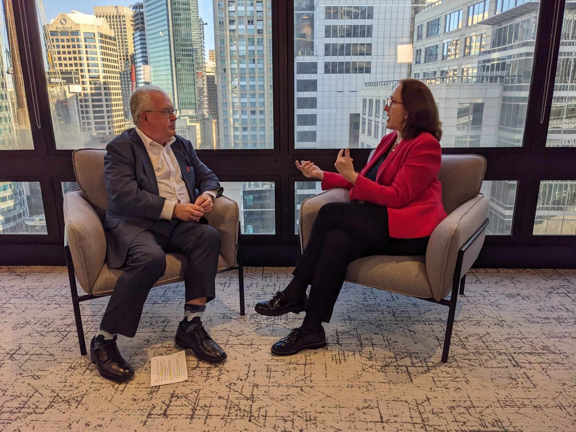 Two people in business attire sit facing each other in armchairs by large windows with a city skyline in the background, engaged in conversation. A sheet of paper lies on the floor near one person.