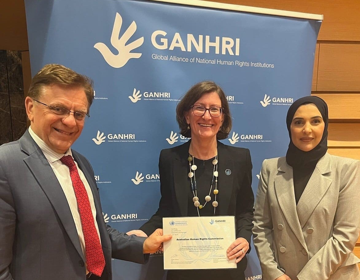 Three people stand in front of a blue GANHRI banner; the person in the center holds a certificate and smiles, while the two others, one in a suit and one in a headscarf, stand on either side, also smiling.