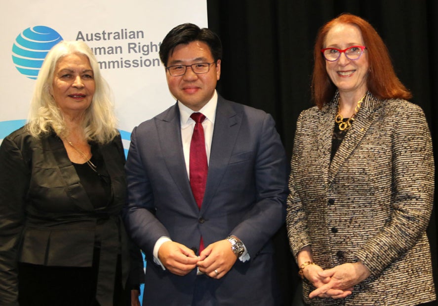 Three people stand together, smiling, in front of an Australian Human Rights Commission banner. The group includes two women, one with white hair, and a man in a blue suit and glasses.