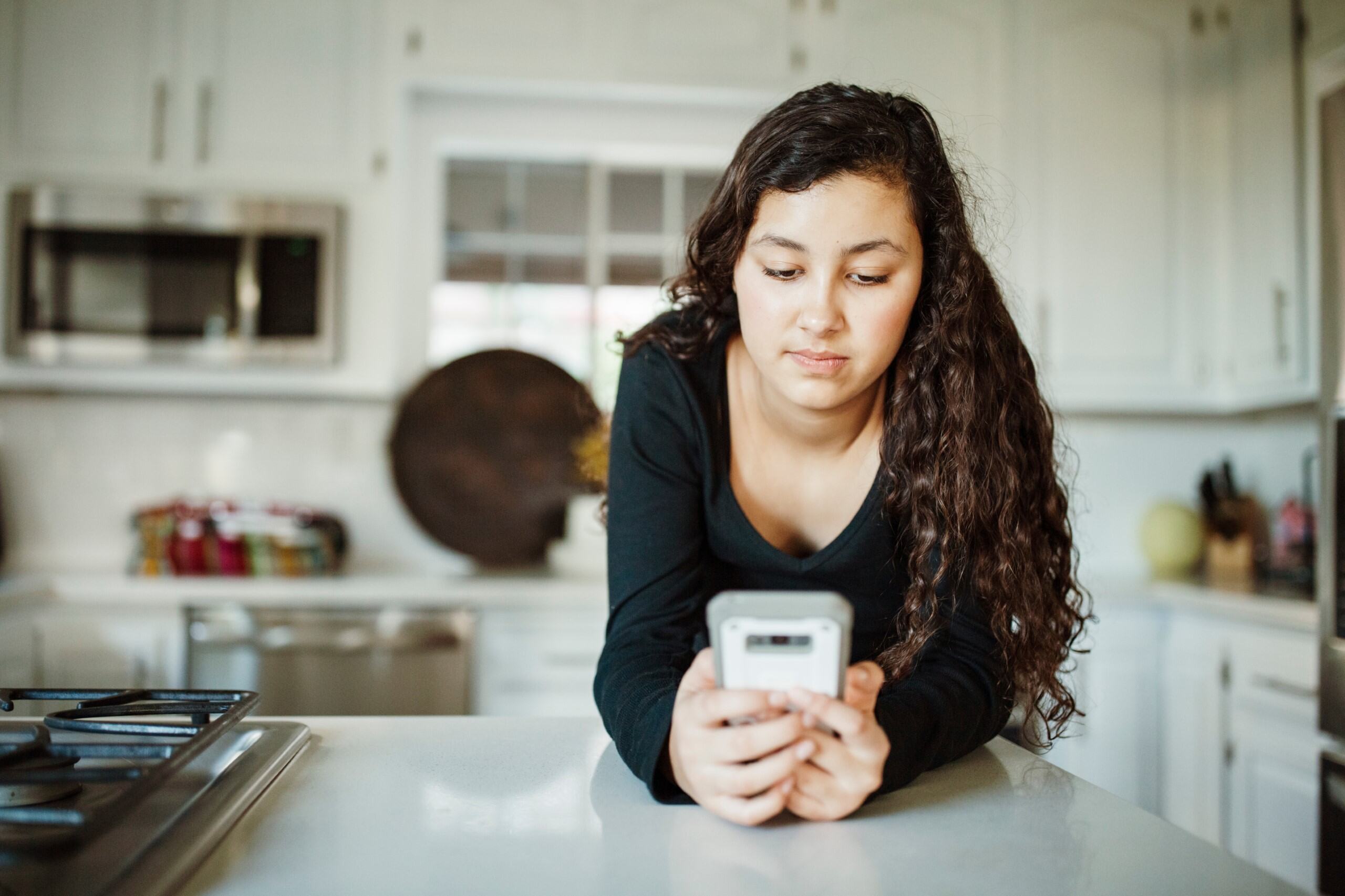 Front view of Hispanic girl, aged around 15, leaning on kitchen counter holding and looking at smart phone.