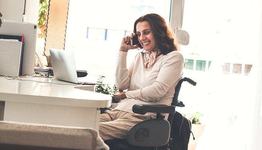 A person in a wheelchair sits at their desk while talking on the phone smiling
