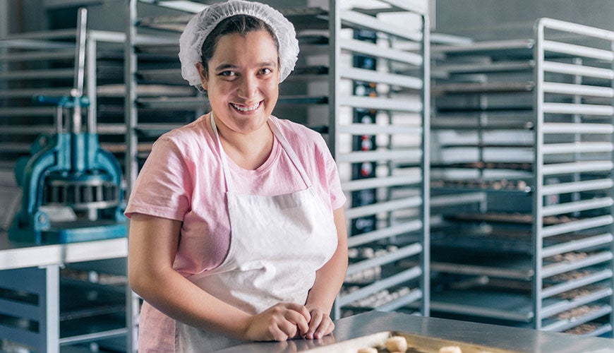 A person with a disability stands, smiling at their workplace in a bakery