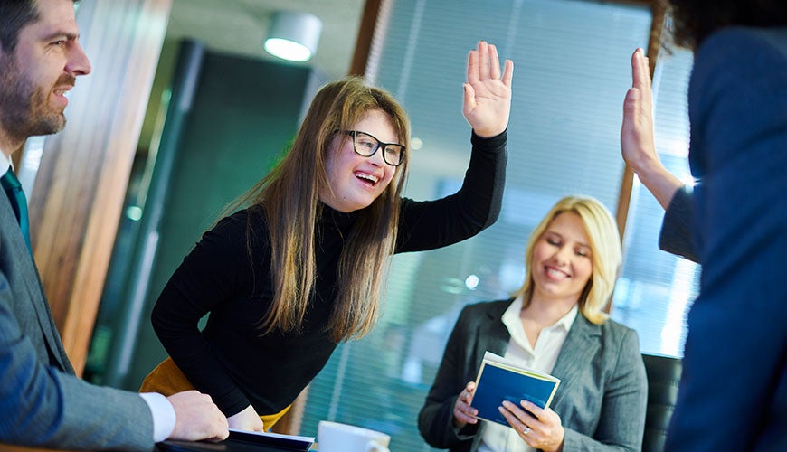 A person with Down Syndrome holds her hand up high-fiving colleagues in an office setting