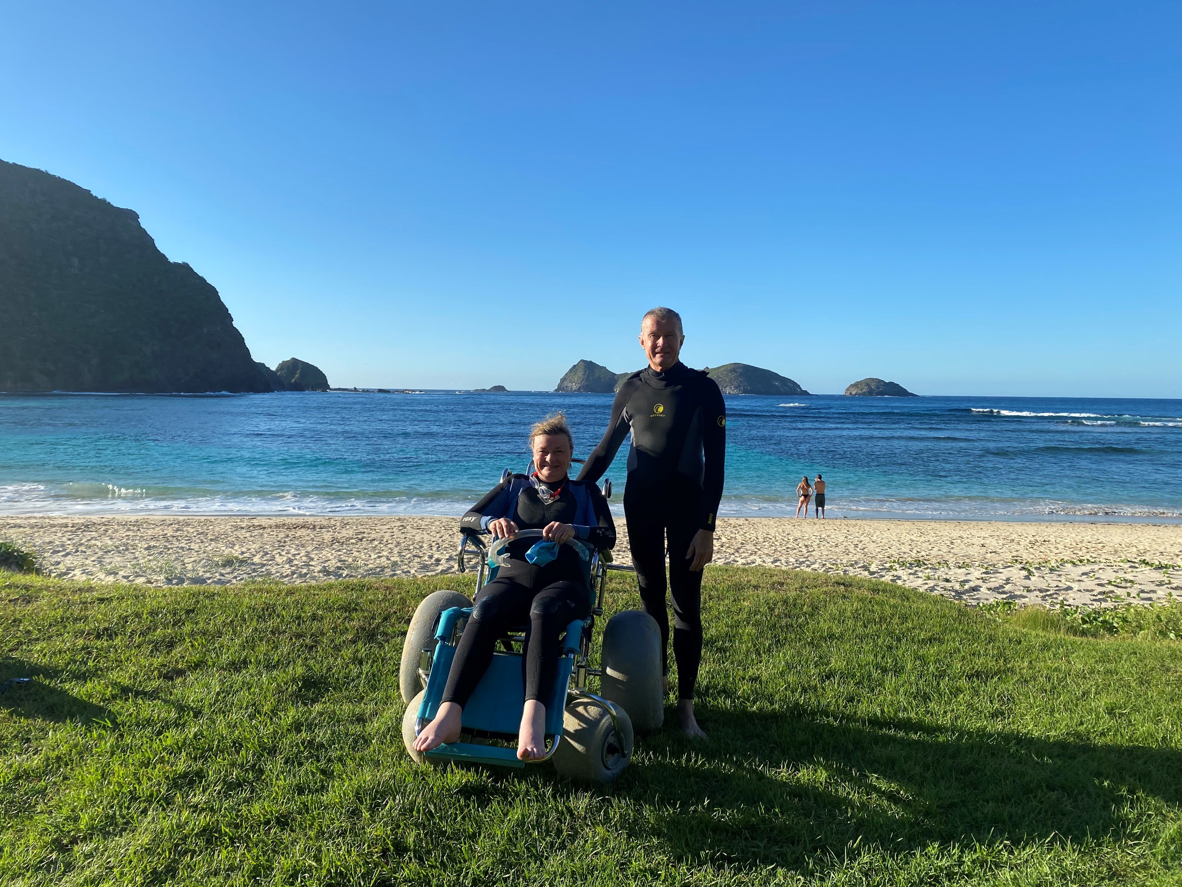 Image of Jane with another person. Jane is sitting in a wheelchair and the man is standing. They are on grass with white sand and blue water behind them. There are mountains behind the water. Both people are smiling. The sky is blue.