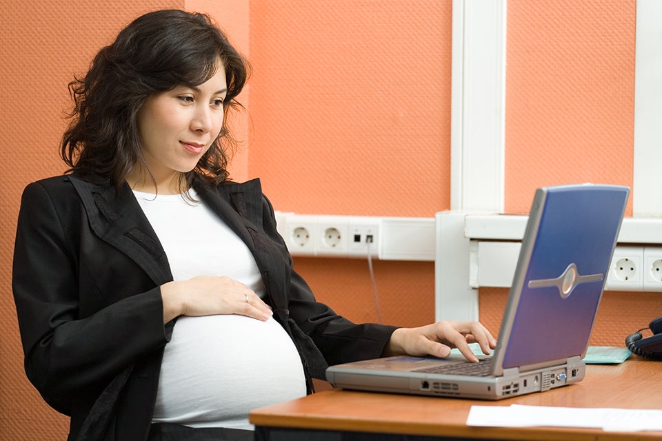 Pregnant lady sitting in front of her laptop with hand over her baby bump