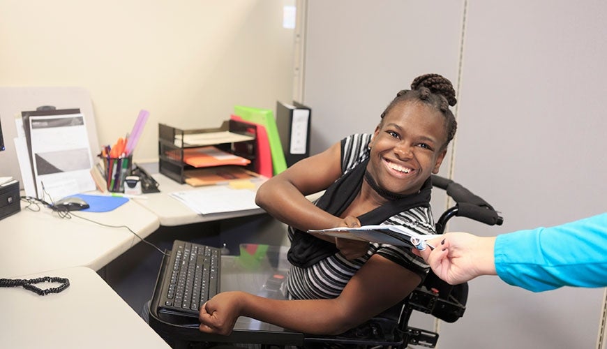 A woman of colour sitting in a wheelchair at her desk with a keyboard positioned in a way that is accessible to her. She is smiling at the camera and accepting some documents from an individual out of frame.