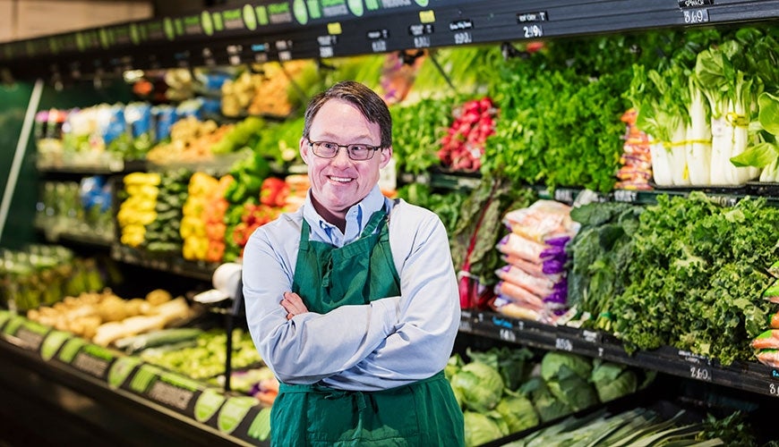 A man working in a grocery store. He wears a buttoned up shirt and a green apron. He appears to have Down Syndrome.