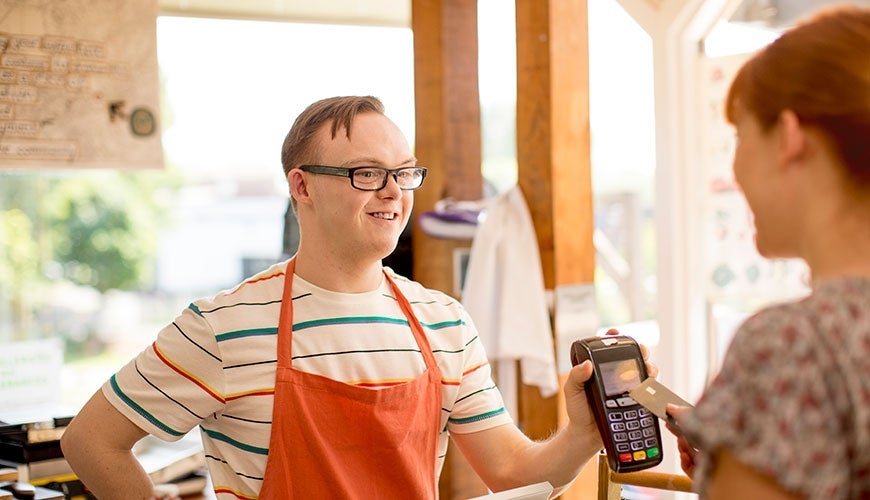 A young man with Down Syndrome is holding up an EFTPOS machine to a customer. He is working in a café. 