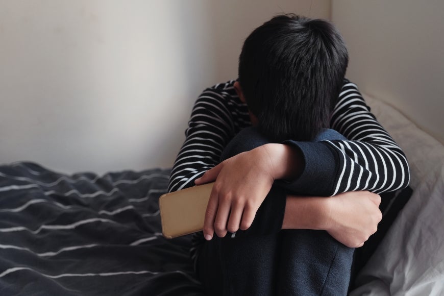 Asian teenager boy hugging his knee in his bedroom with smartphone.