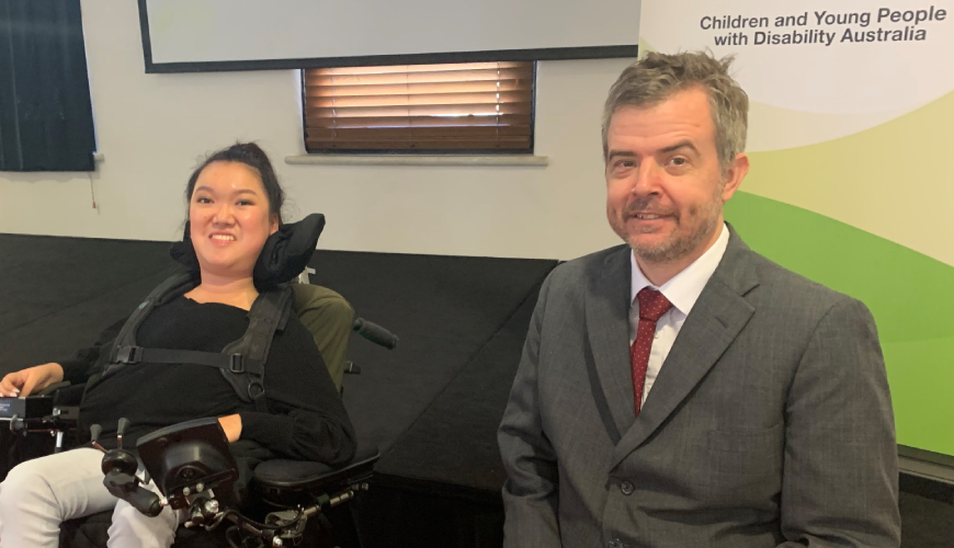 Ben Gauntlett and Melanie Tran, who use wheelchairs, in front of a small stage. Both are smiling and looking at the camera.