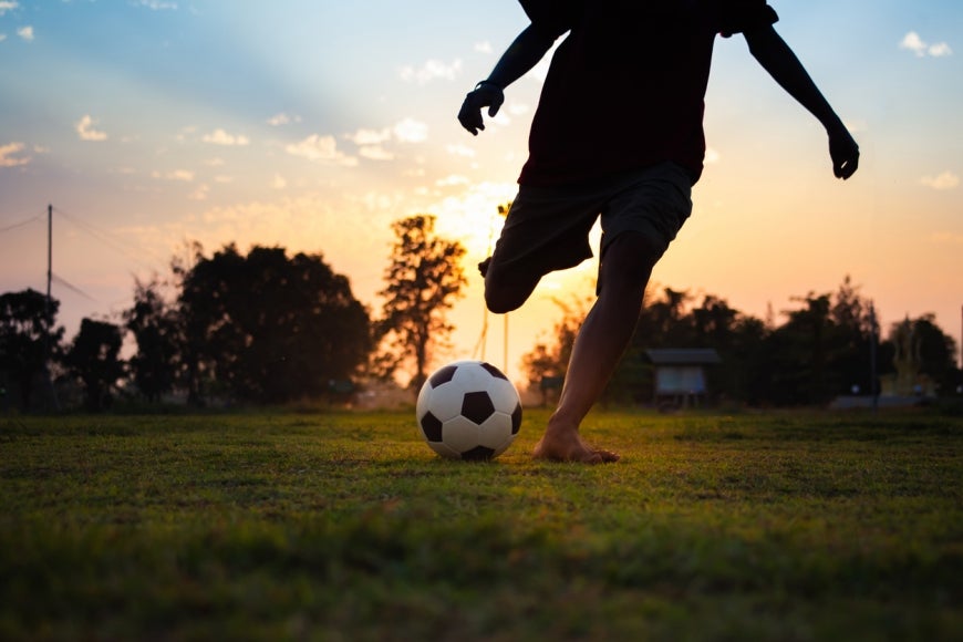 A young person plays soccer on an open field alone, with the sun setting behind them.