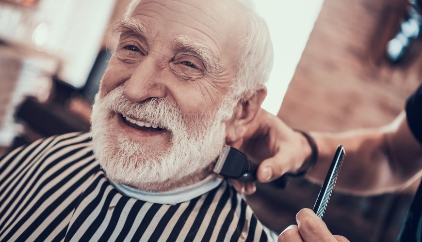 Smiling grey haired man while his beard is trimmed with a machine.
