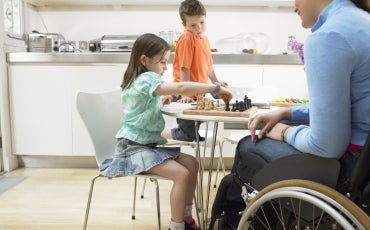 A woman using a wheelchair is at a table with two young children at home playing a game.