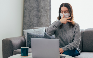 A young woman sits on a sofa looking at a laptop, using sign language and wearing a mask.