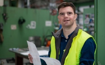 A man with Down Syndrome in an industrial working environment holding some paperwork and wearing a safety vest.