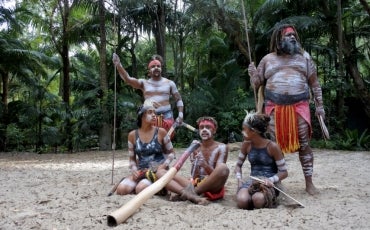 First Nations children in traditional dress, surrounded by elders, sitting on the sand holding traditional instruments like didgeridoo