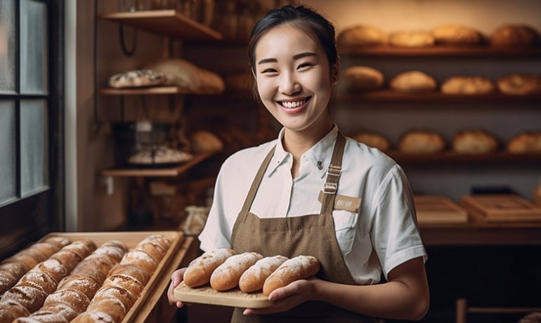 Women in a bakery smiling and holding a tray of bread rolls