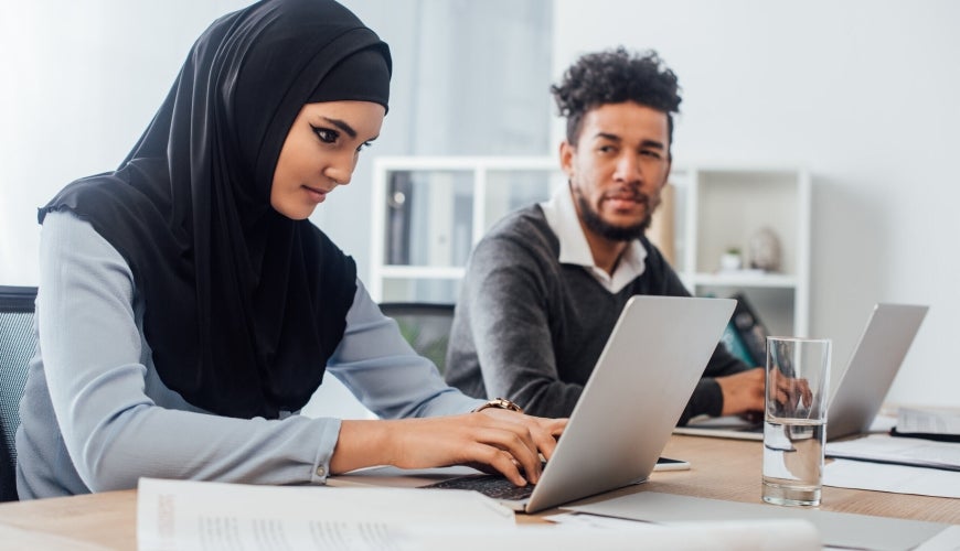 "alt="Muslim business woman working on laptop with man in background watching."