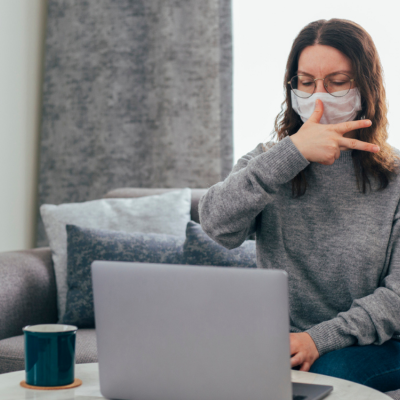 A young woman sits on a sofa looking at a laptop, using sign language and wearing a mask.