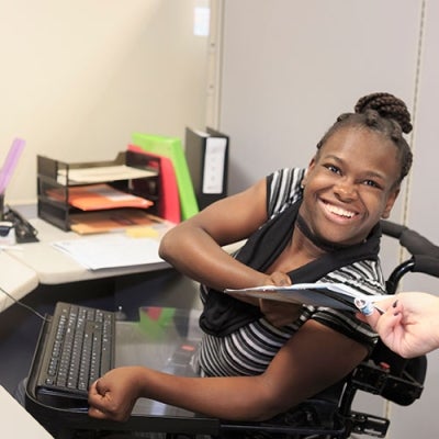 A woman of colour sitting in a wheelchair at her desk with a keyboard positioned in a way that is accessible to her. She is smiling at the camera and accepting some documents from an individual out of frame.