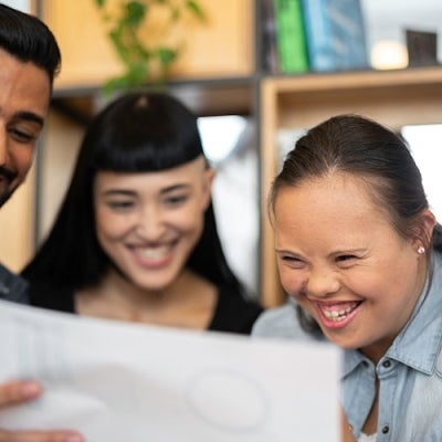 Three people smiling and looking at a work plan together. From left to right: There is a man with dark skin and an earring, a woman with long black hair and another woman also with long dark hair, which is pulled back, who appears to have Down Syndrome.