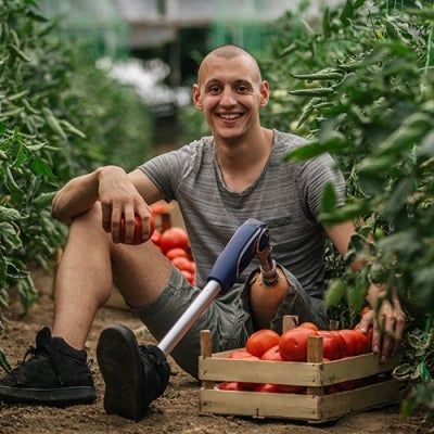 A young man with a prosthetic leg is sitting on the ground between rows of tomato plants. He is picking tomatoes and smiling