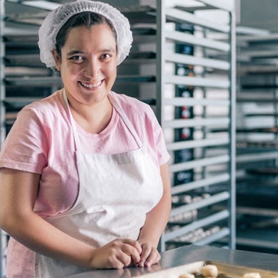 A woman wearing an apron and hairnet is smiling in an industrial bakery kitchen. She has a tray of pastries in front of her.