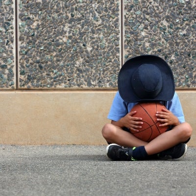School boy sitting cross legged hiding face in a basketball.
