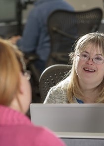 A person with Down Syndrome smiles and looks over the top of their work laptop to chat with a colleague.