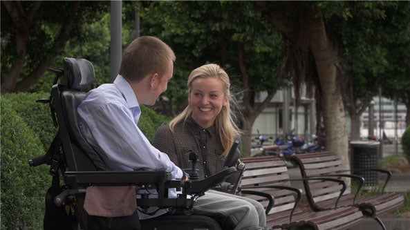 still image: man on wheelchair and woman on bench facing to each other, smiling